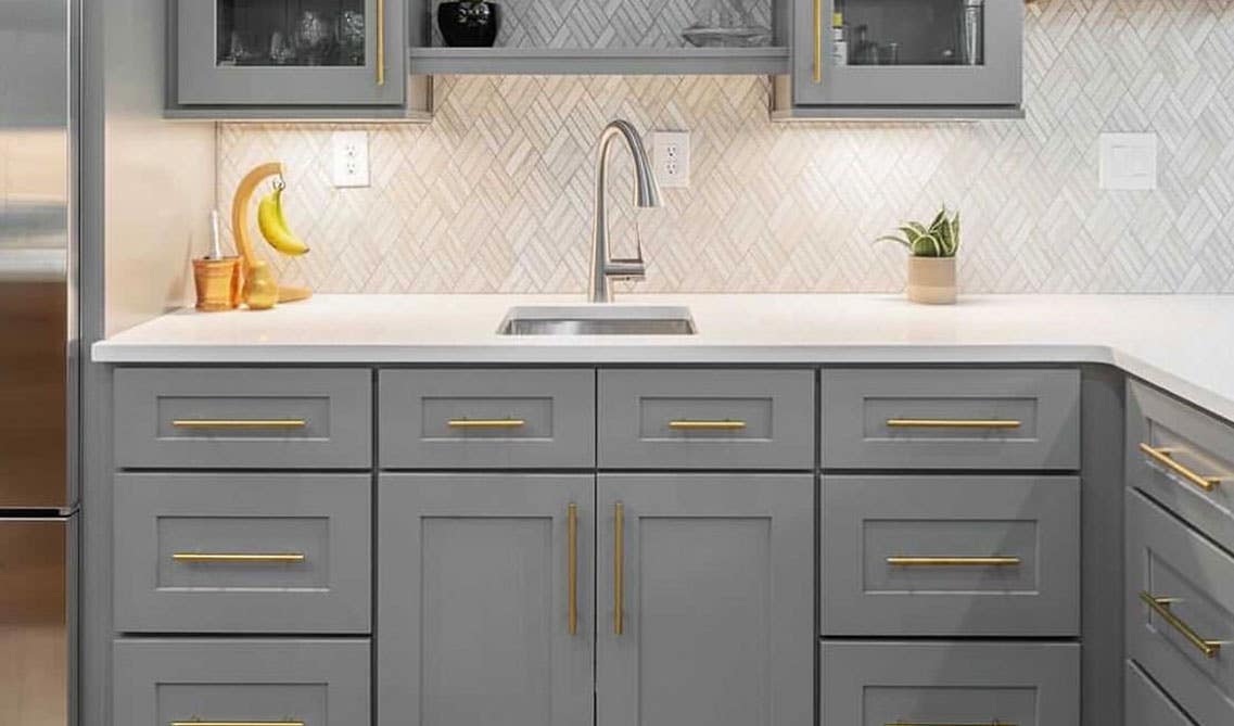 Kitchen gray cabinets with gold pulls, floating shelf above stainless steel sink and white quartz countertop