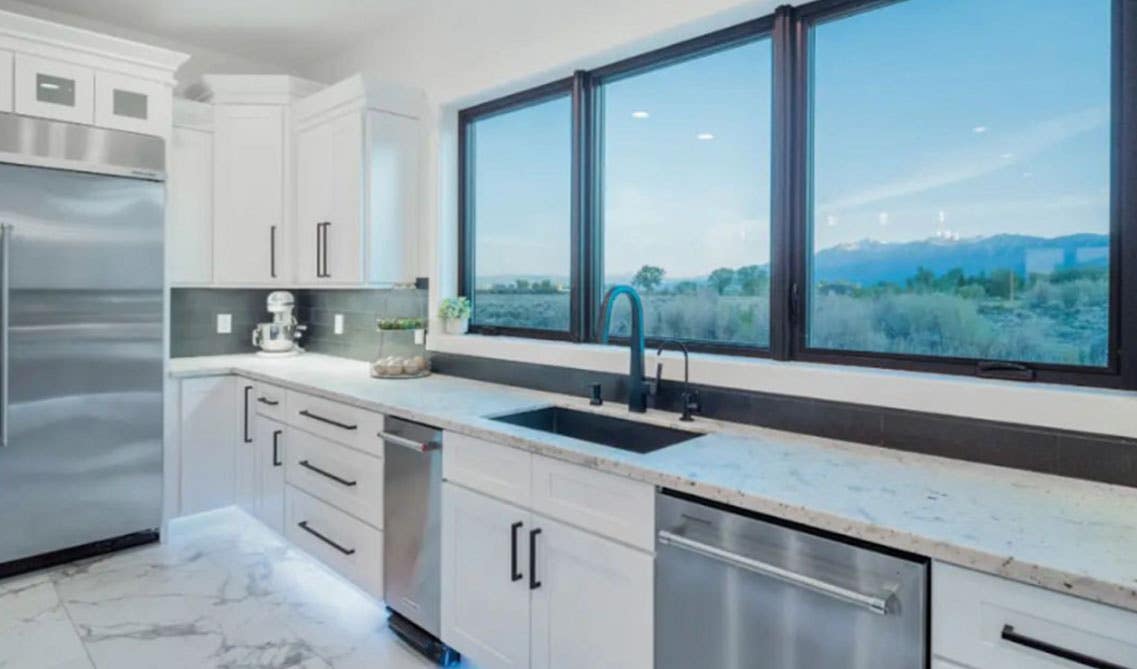 Modern kitchen design with white shaker cabinets, quartz countertops, stainless steel appliances and large window above sink