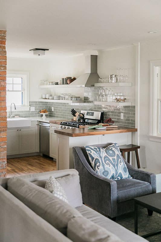 small kitchen with white shaker cabinets and glossy gray subway tile backsplash. floating shelves house dishware and a wooden butcher block countertop is adjacent to a farmhouse sink