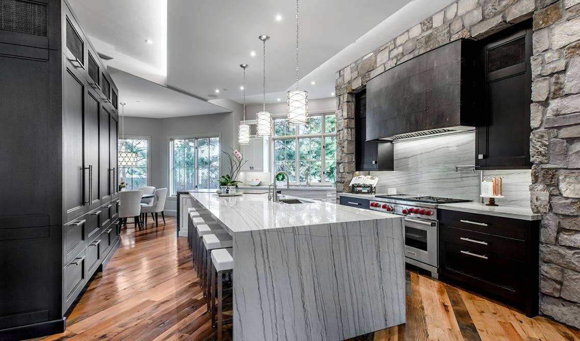 unique rustic kitchen design with hardwood floors, a natural stone accent wall, dark grey cabinets, and a waterfall marble island