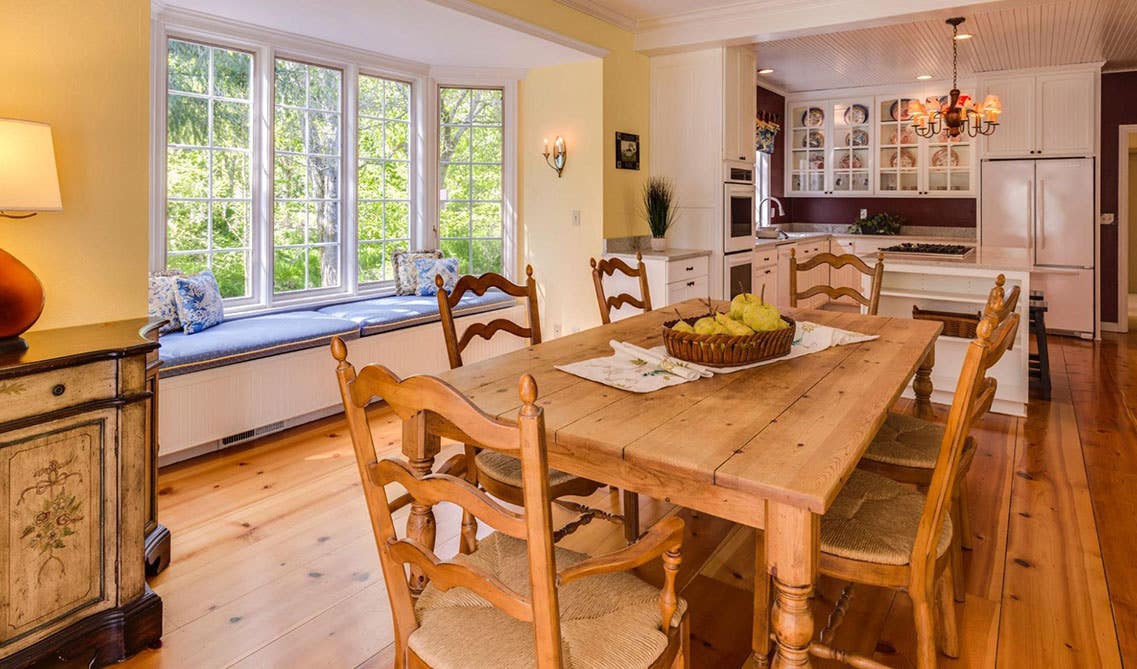 Traditional kitchen with white cabinets and natural wood dining table, large window sitting area and hardwood flooring