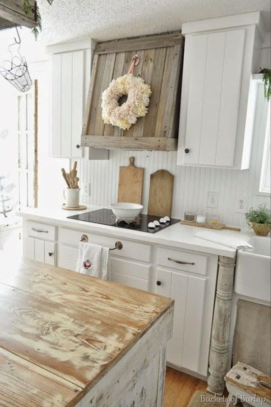 Distressed vintage farmhouse kitchen with white panel cabinets, wood range hood, butcher block island, and shiplap backsplash