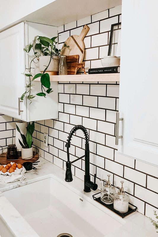 modern farmhouse kitchen with white subway tile backsplash and black grout. a floating shelf sits between two white cabinets, above a farmhouse sink with black hardware