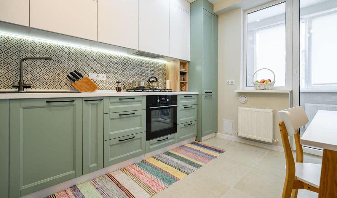 two toned green and white kitchen cabinets with black hardware, pattern wallpaper as backsplash, tile flooring with multicolor rug