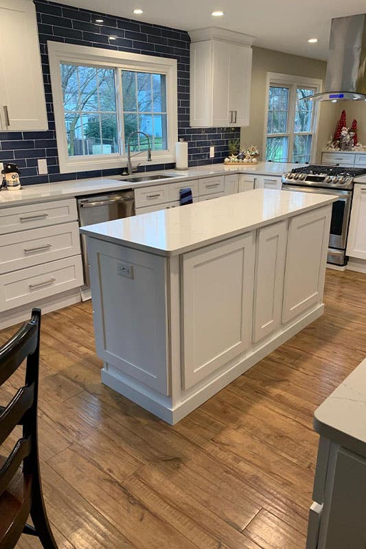 Modern kitchen features white shaker cabinets, small island, white marble countertops, navy blue backsplash and stainless steel appliances