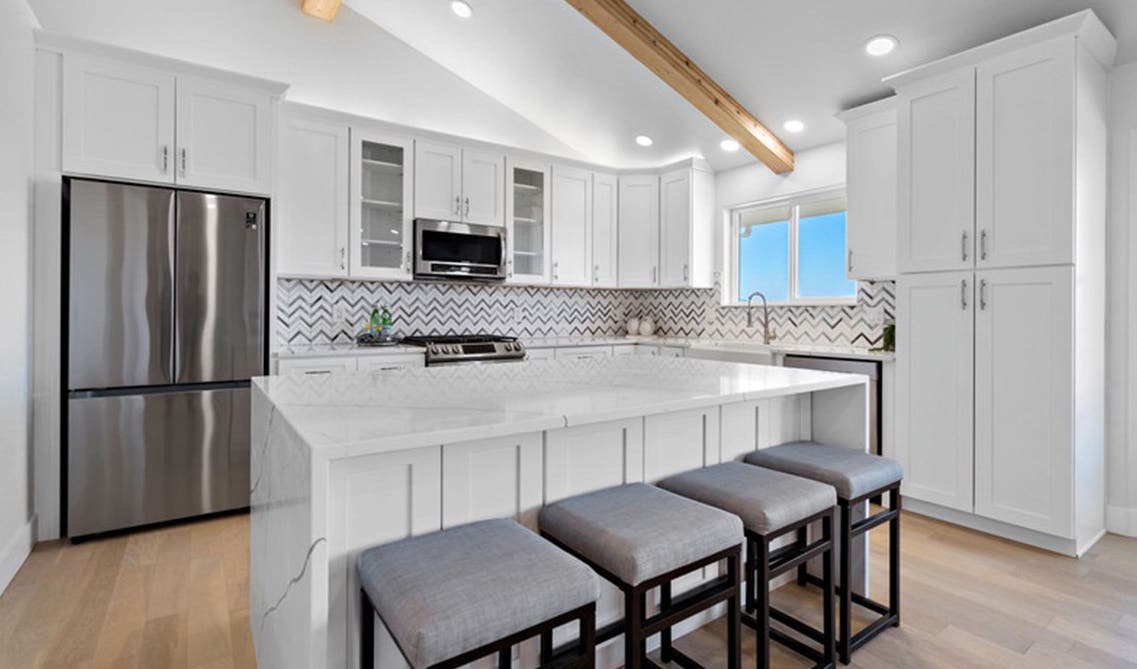 White kitchen with shaker cabinets, large island with waterfall countertop, 4 stool chair, and stainless steel appliances