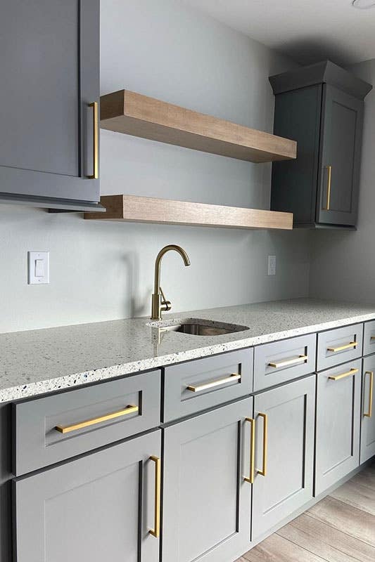 Small wet bar are with gray cabinets with gold pulls and bar sink with marble countertops, open shelving