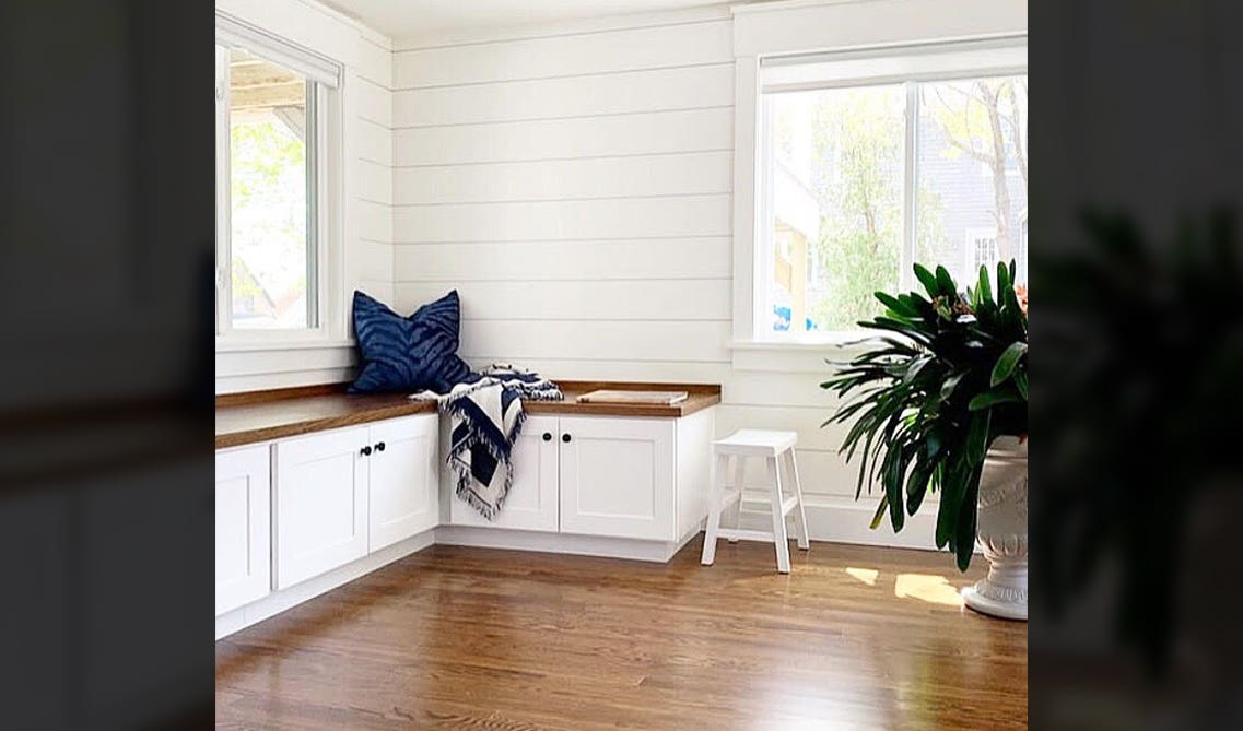 Mudroom idea with white base cabinet with sitting bench, blue pillow, white stool, big flower vase, 2 large windows