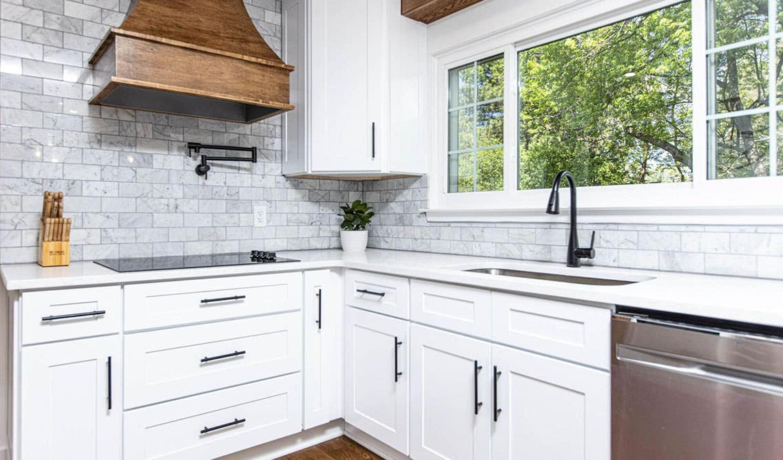 white shaker elite cabinets with black hardware, white countertop, wood rang hood and large window